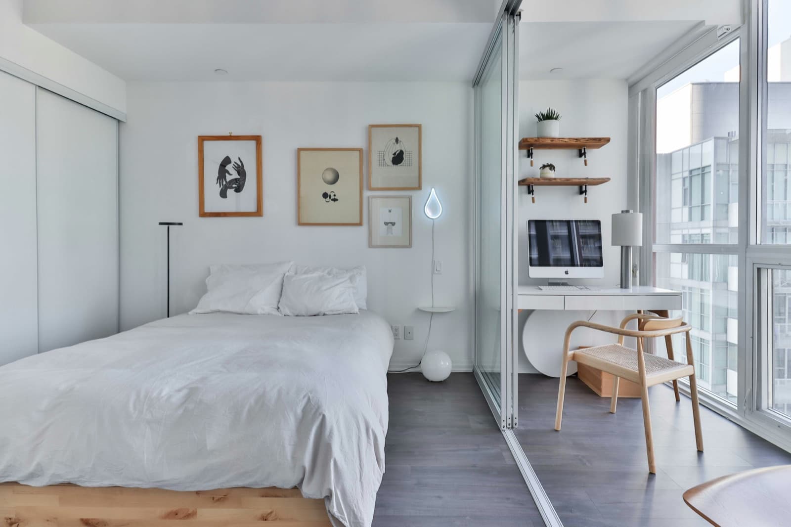 A small condo bedroom painted in a soft white, with a glass partition and a neighbouring tower visible through the window